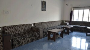 A spacious living room with a large striped sofa, a small table, and a bench. There is a window with curtains and a painting on the wall. - Hotel Joshi (Siddharthanagar)