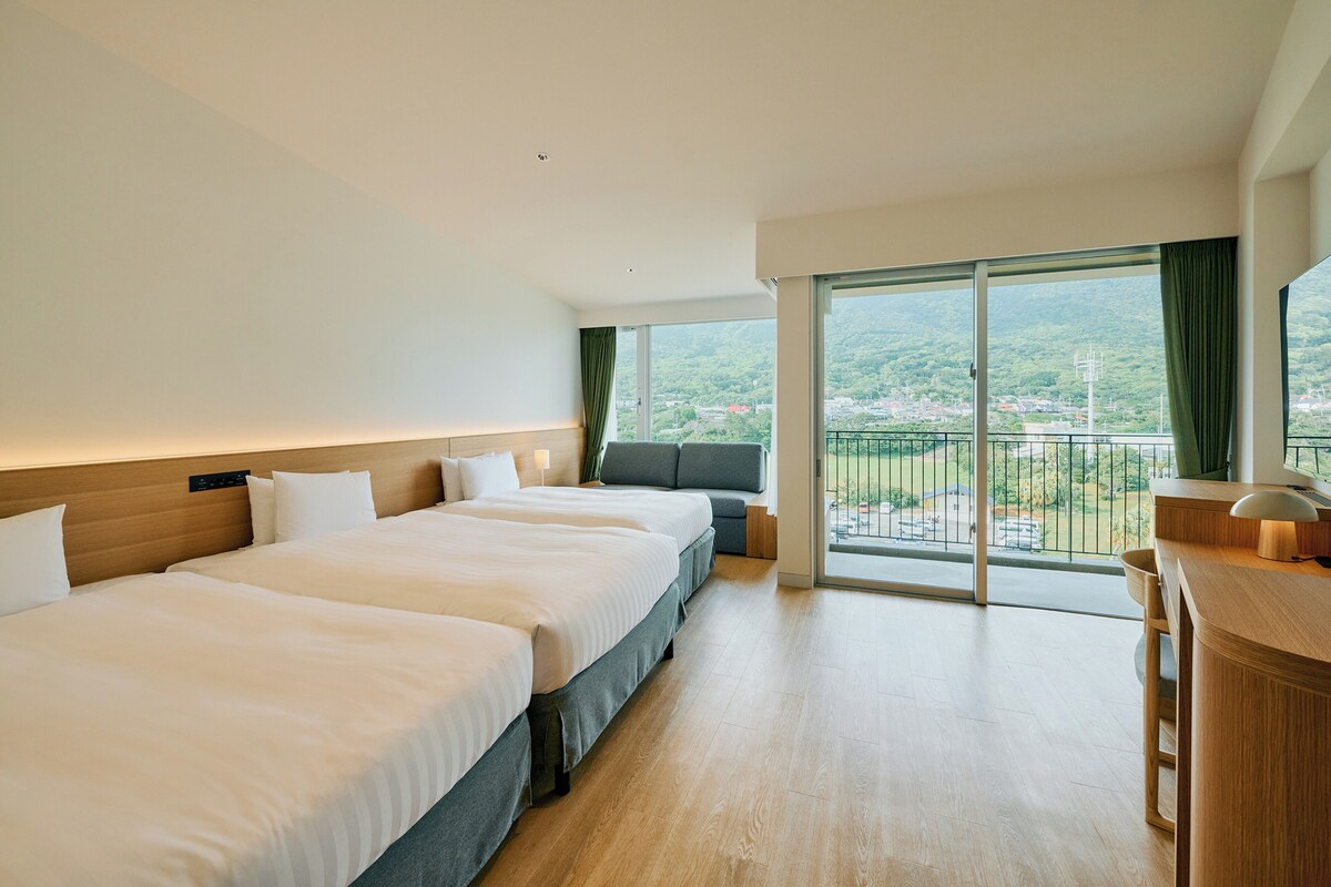 Ein Hotelzimmer mit zwei Betten, einem Schreibtisch und einem Balkon mit Blick auf eine Stadt und Berge.