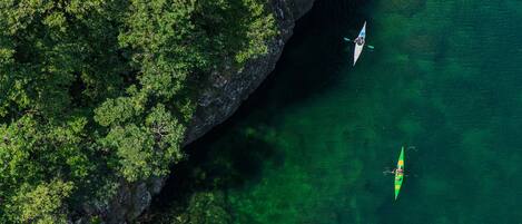 An aerial view of a green kayak on clear water near a rocky cliff with dense green foliage.