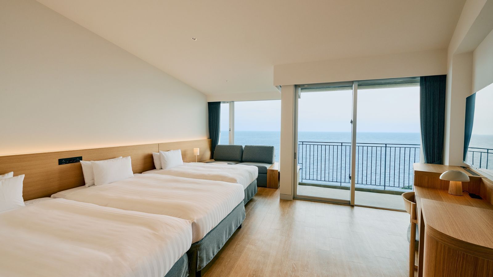 Ein Hotelzimmer mit drei Betten, einem Schreibtisch und einem Balkon mit Meerblick.