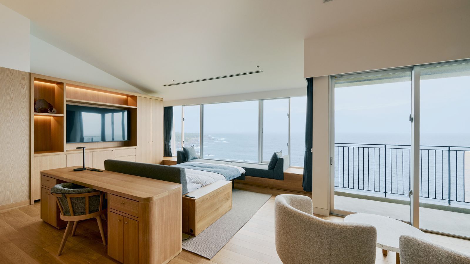 Ein modernes Schlafzimmer mit Bett, Schreibtisch, Fernseher und einem Balkon mit Meerblick.
