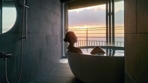 Une salle de bain moderne avec une grande baignoire, une douche et une vue sur le coucher de soleil sur l’océan.