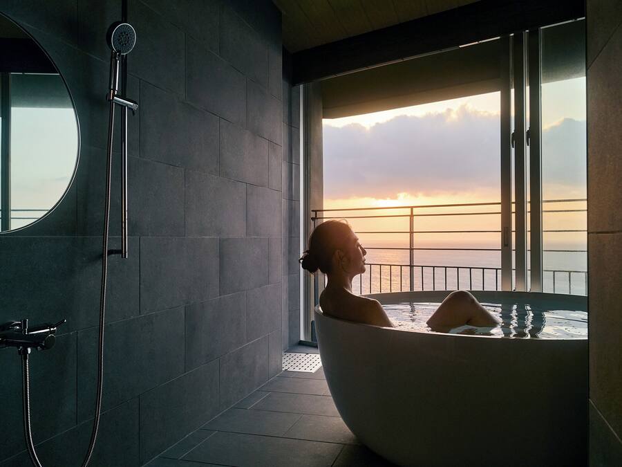 A modern bathroom with a large bathtub, a showerhead, and a view of the sunset over the ocean.
