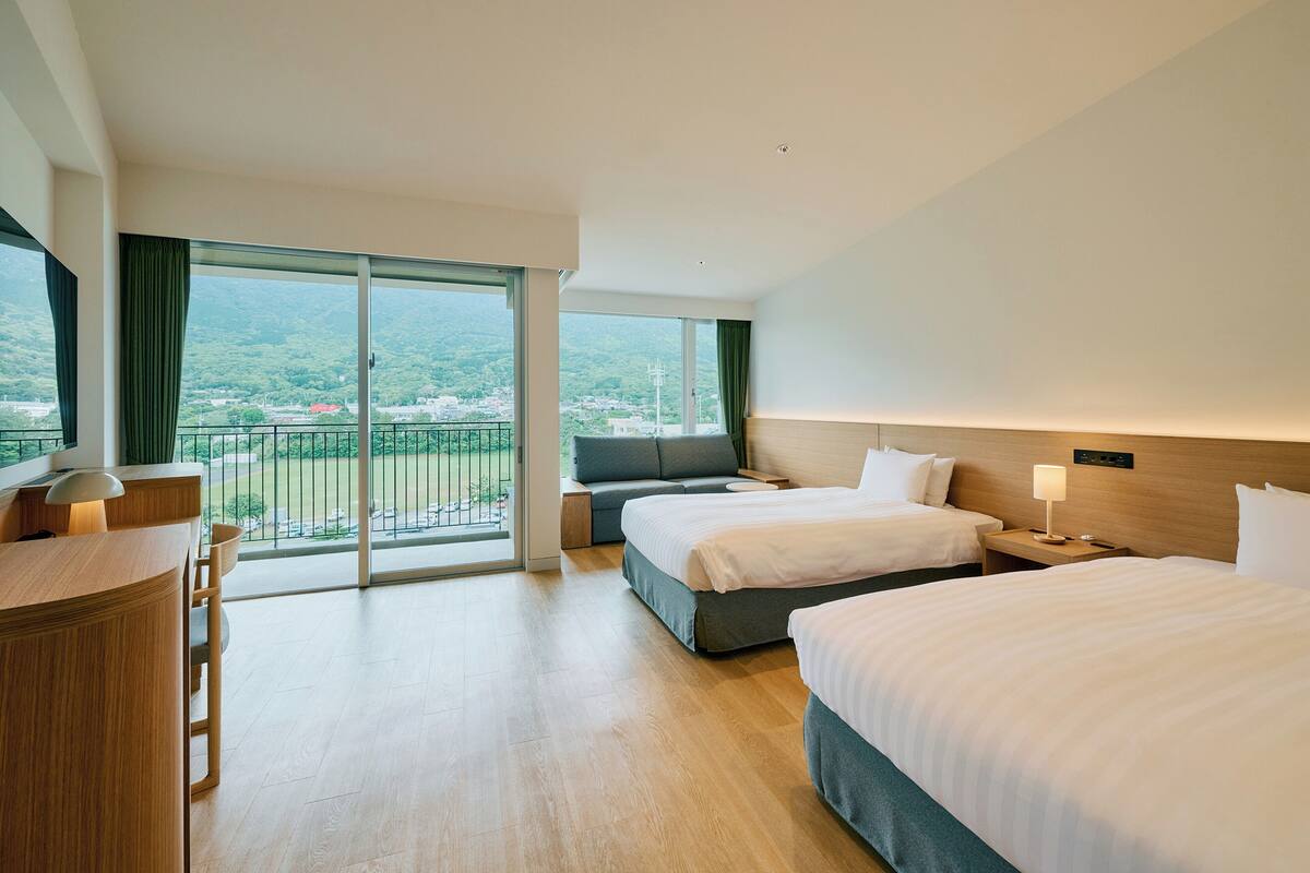 Een hotelkamer met twee bedden, een bureau, een televisie en een balkon met uitzicht op de stad en de bergen.