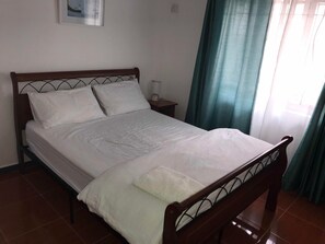 A neatly made bed with white linens and a wooden headboard in a room with a tiled floor and a green curtain. - H&K Villa (Blue Bay)