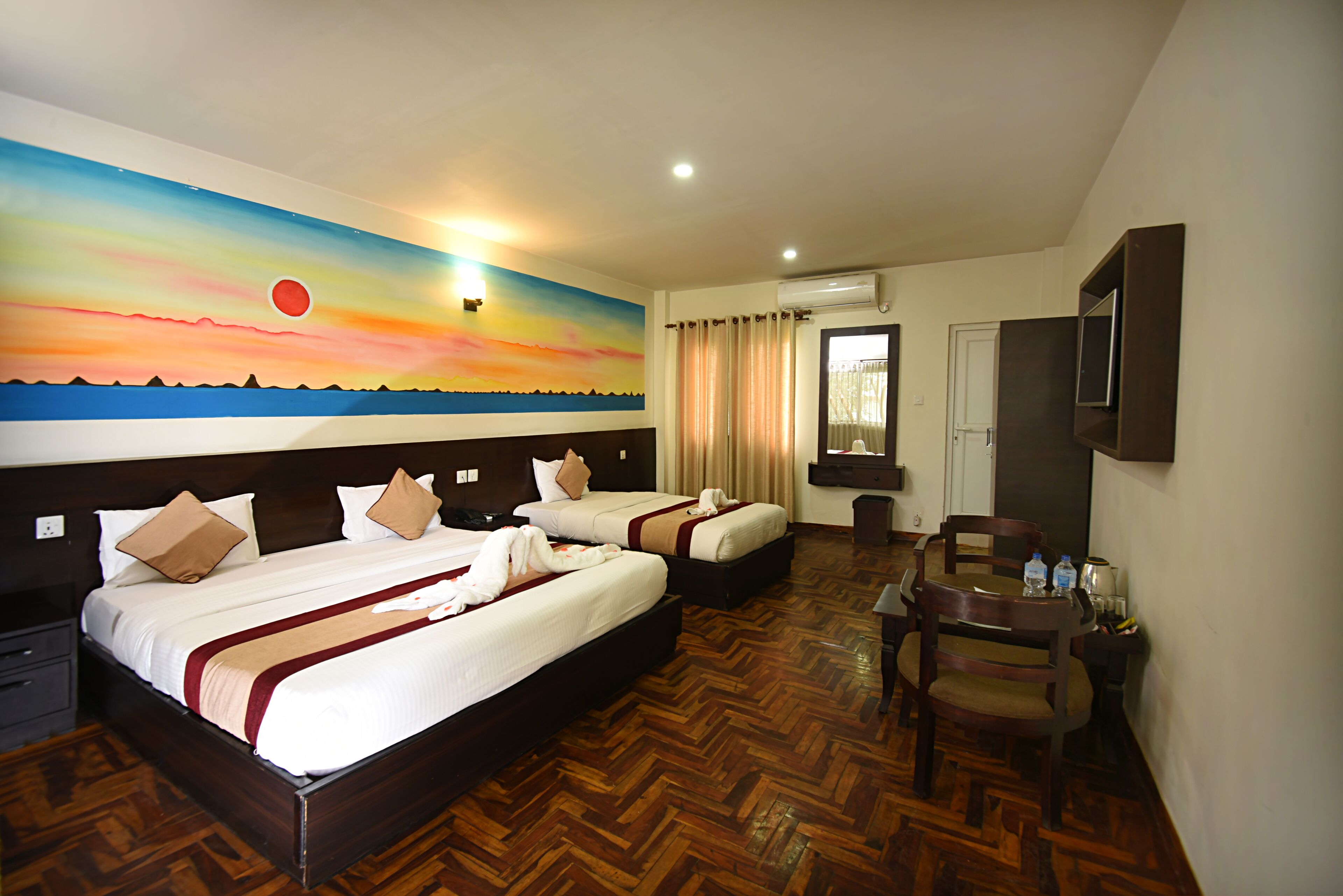 A hotel room with two beds, a wooden herringbone floor, a painting of a sunset, a desk with chairs, and an air conditioning unit.