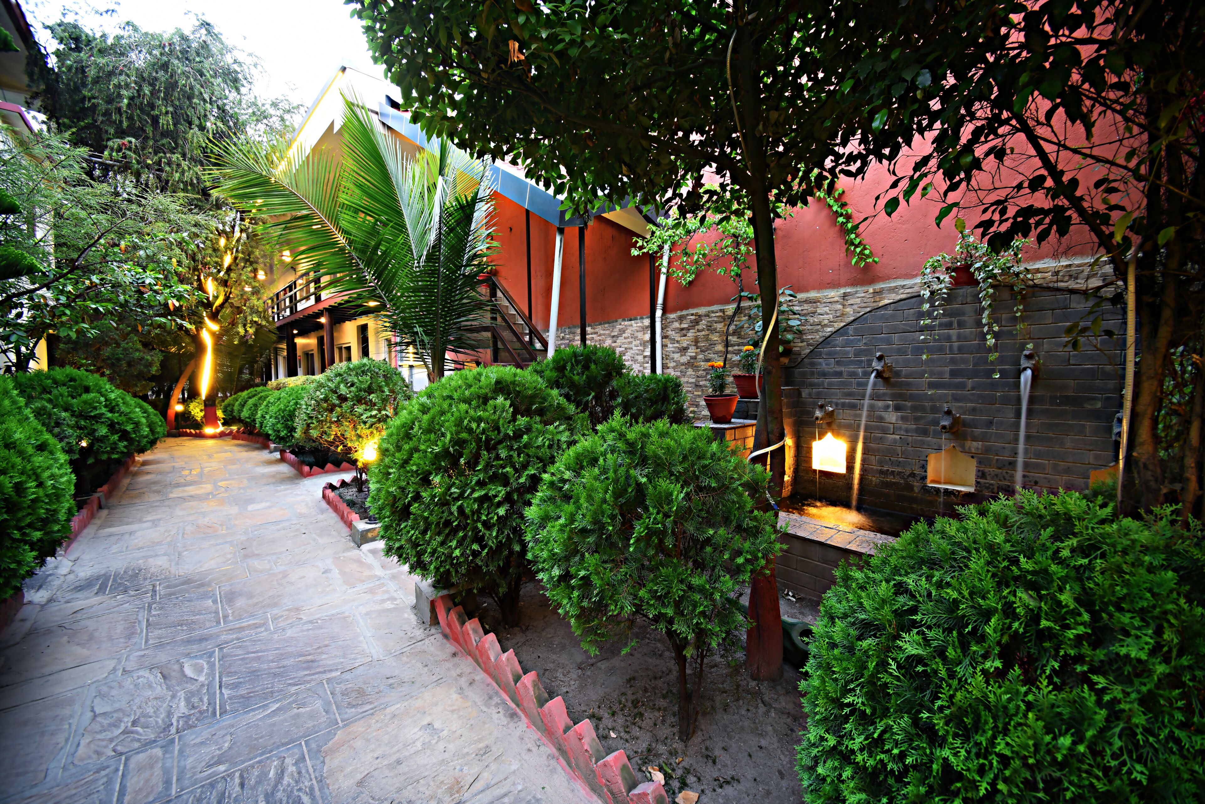 A stone pathway flanked by well-maintained bushes and a brick wall with a water feature.