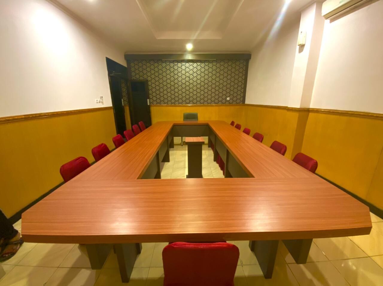 A conference room with a long wooden table, red chairs, and a patterned wall.