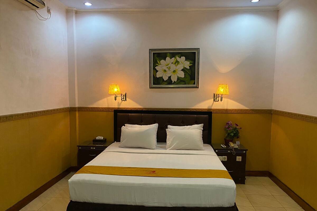 Een hotelkamer met een groot bed, twee bedlampen, een nachtkastje, een bloemistisch kunstwerk in een lijst en een geel accentmuur.