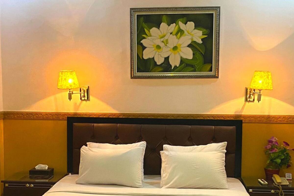 Een hotelkamer met een bed, twee bedlampen, een nachtkastje, een lijst met bloemen en een logo boven het bed.