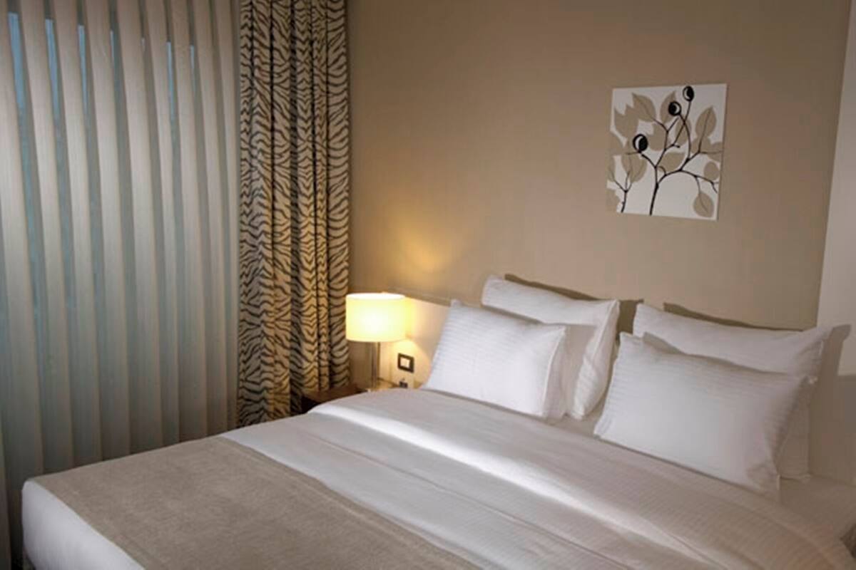 A hotel room with a bed, curtains, a wall-mounted lamp, and a painting.