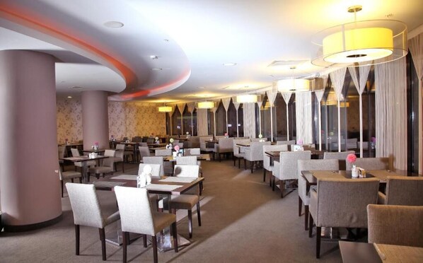 Un restaurante espacioso con numerosas mesas y sillas, amplias ventanas con cortinas e iluminación moderna.