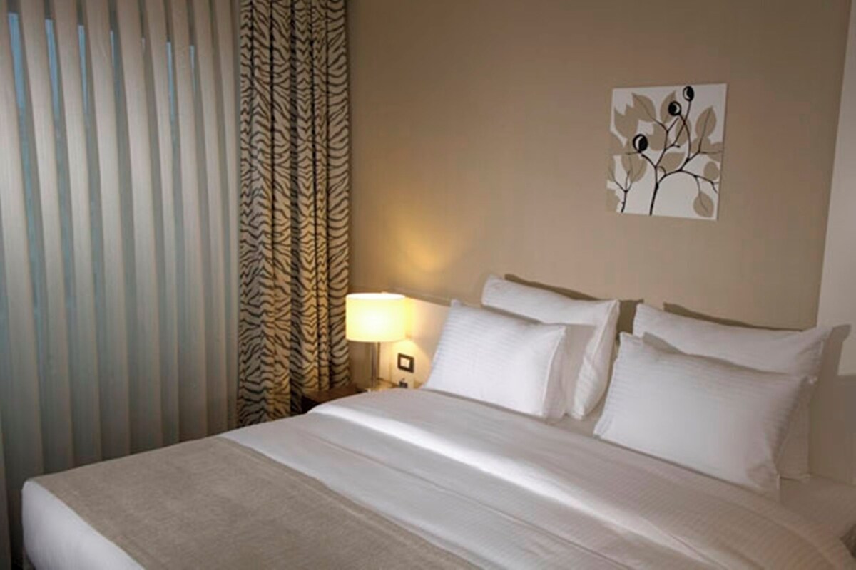Una habitación de hotel con cama, cortinas, una lámpara de pared y un cuadro.