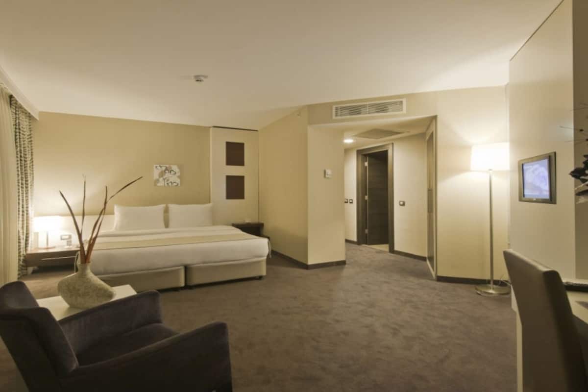 A hotel room with a bed, a desk, a chair, a lamp, and a vase with branches.