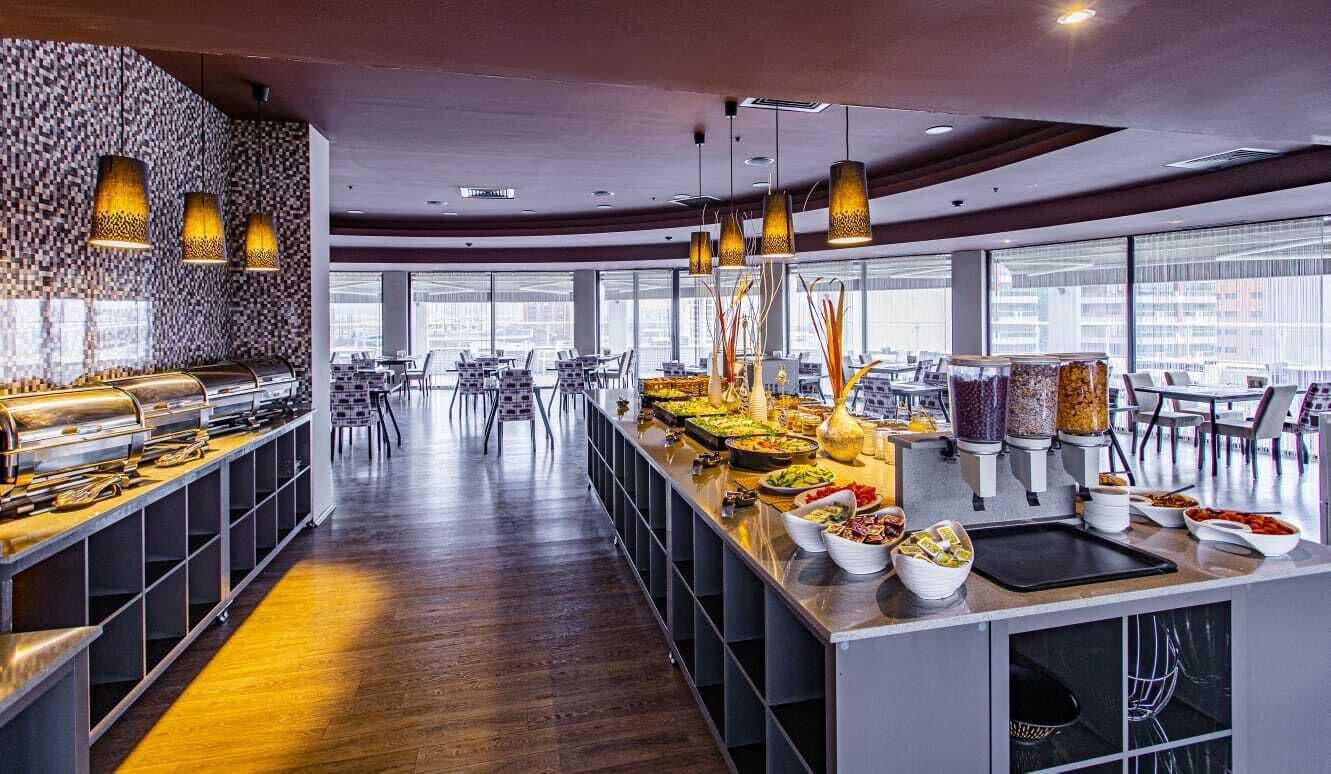 A modern buffet setup with various food stations and seating areas, large windows offering views of the city, and contemporary lighting fixtures.