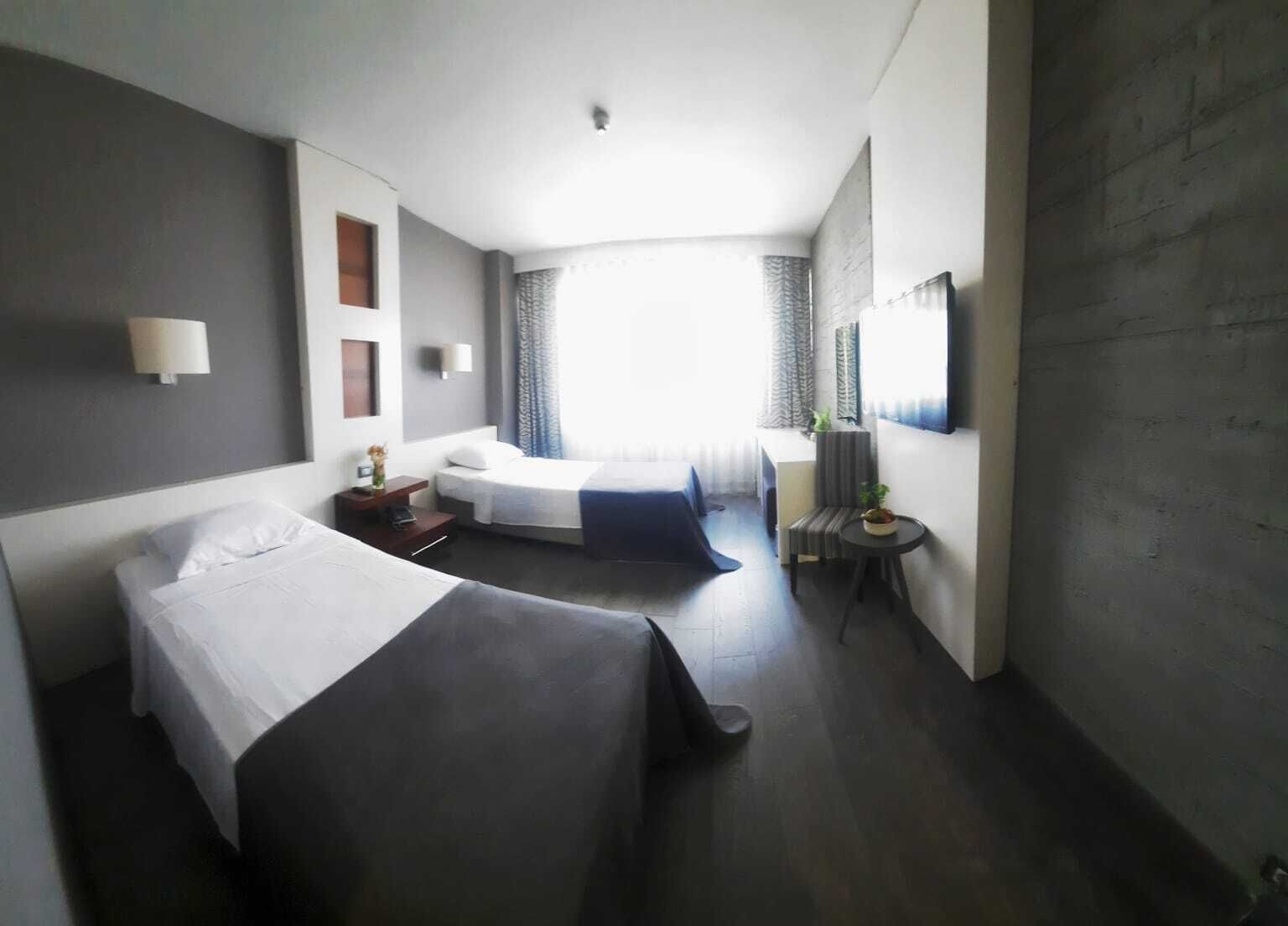 A hotel room with two beds, a small table, a chair, a TV, and a window with curtains.