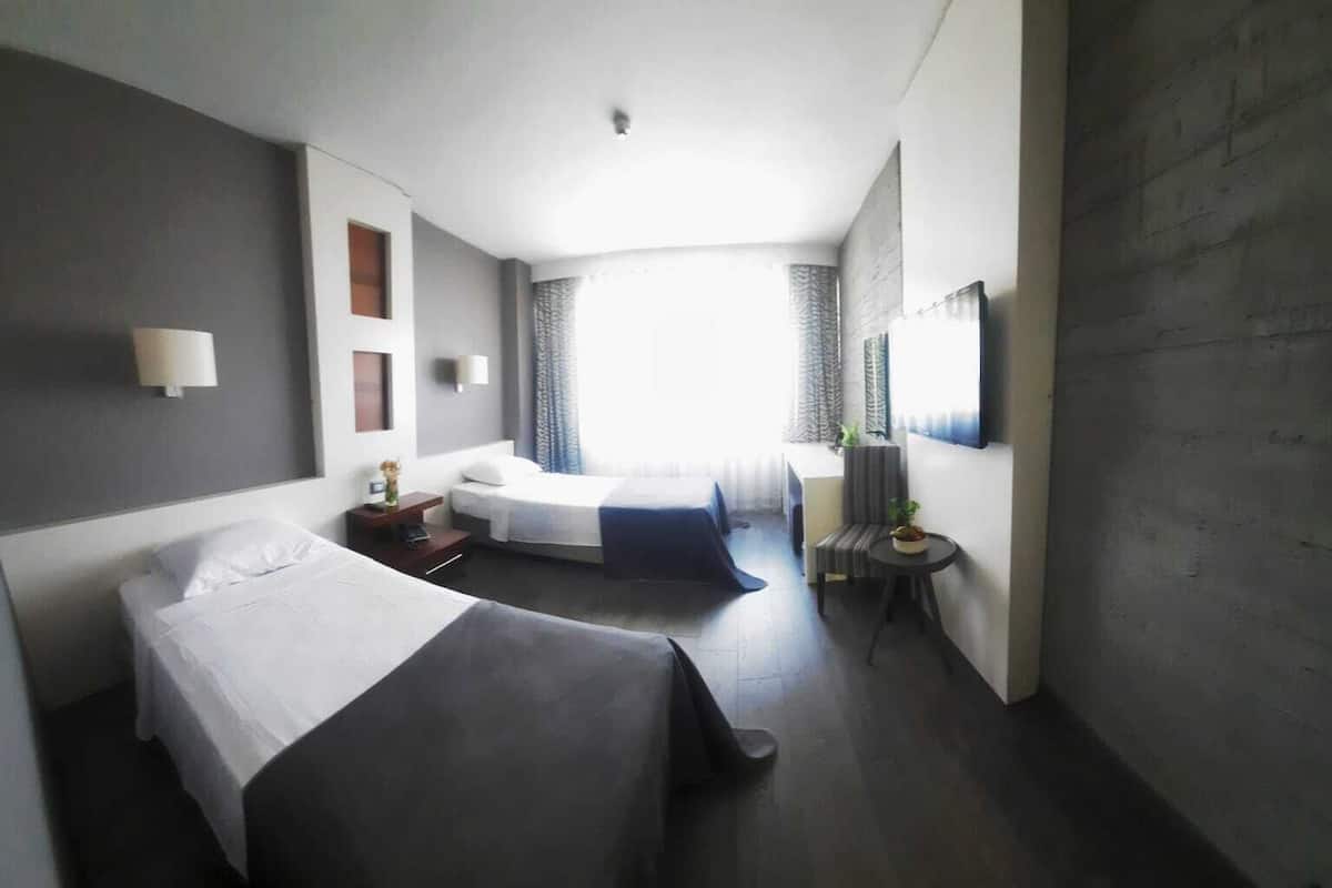 A hotel room with two beds, a small table, a chair, a TV, and a window with curtains.