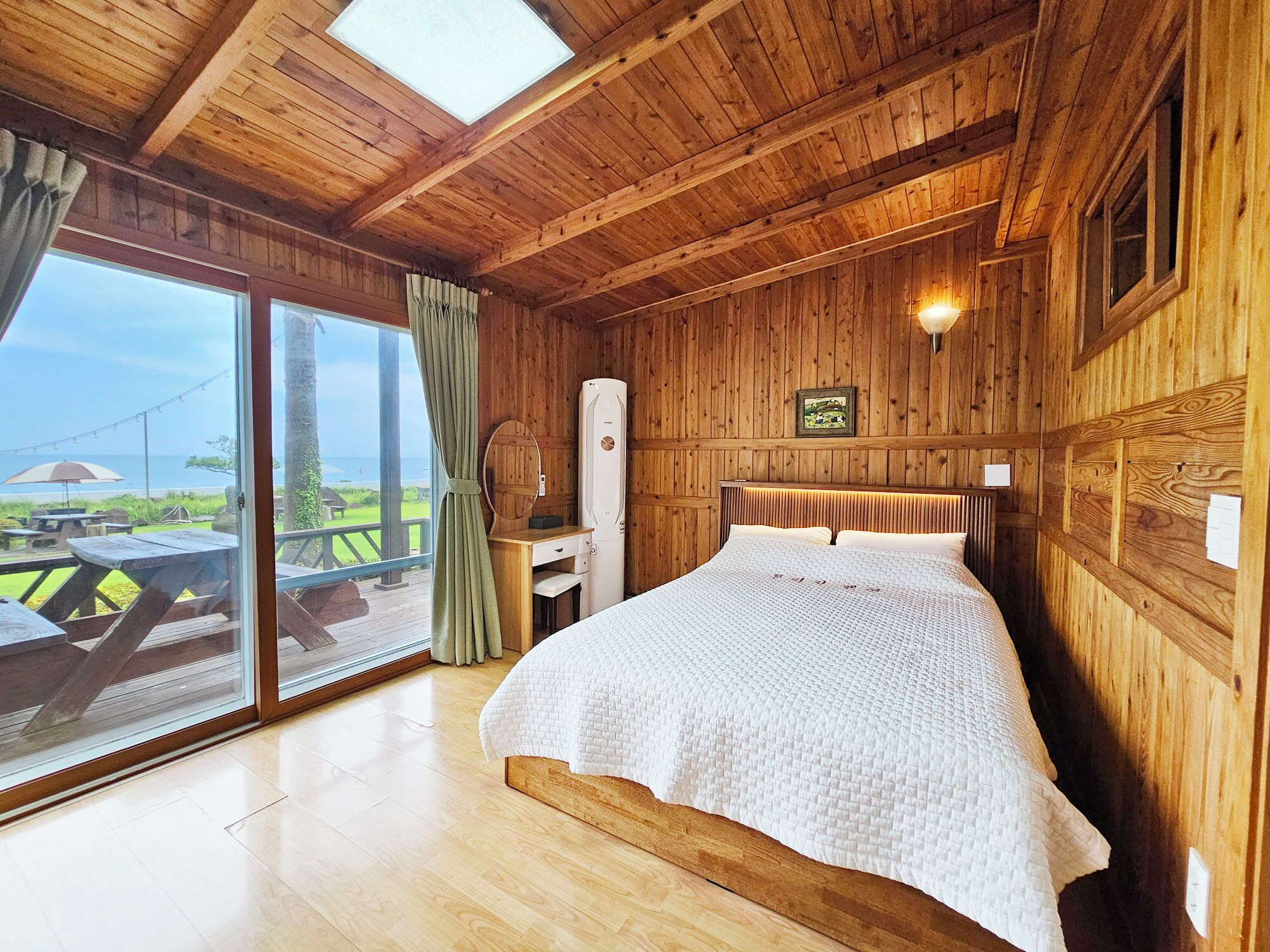 A wooden cabin room with a bed, a desk, and a view of the beach.