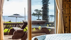 Ett rum med ett stort fönster som vetter ut mot en terrass med picknickbord och parasoller, en trädörr och en säng med ett blått täcke.