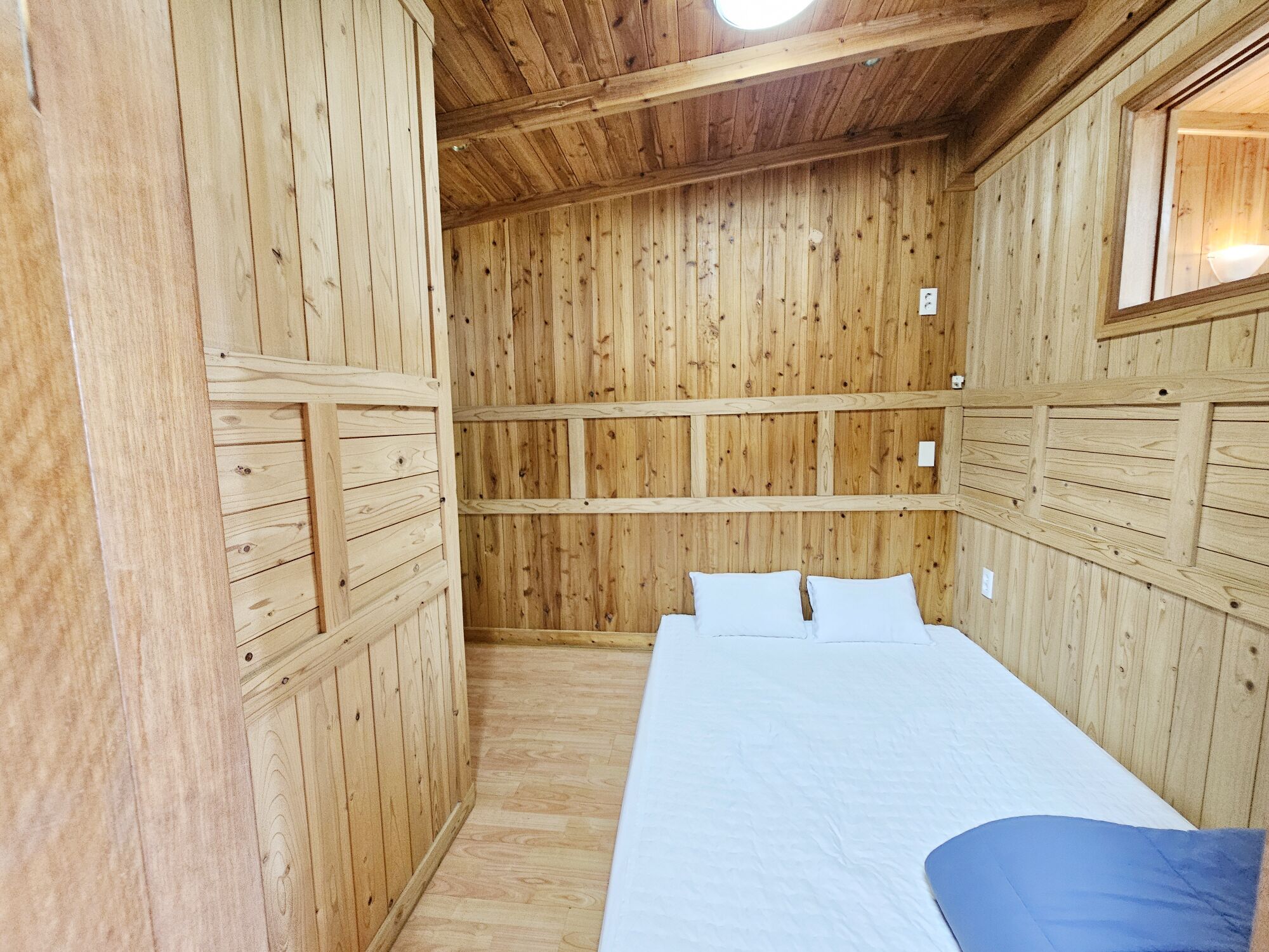 A wooden-paneled room with a bed, a window, and a ceiling light.