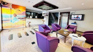Eine moderne Lobby mit violetten Sitzgelegenheiten, einer Rezeption, einem Fernseher und einem Wandgemälde von einem Mädchen am Meer.