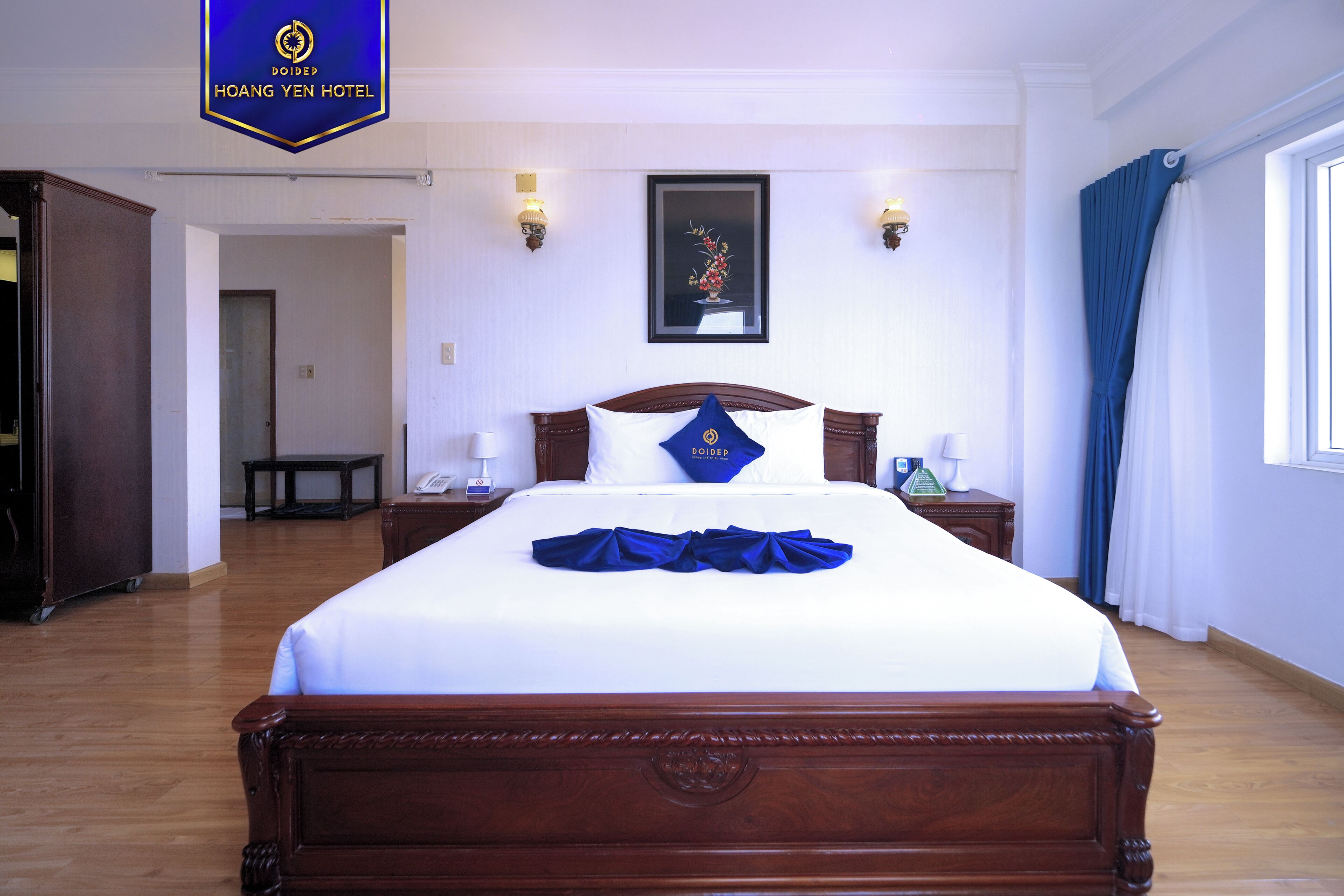 A hotel room with a large bed, wooden headboard, two bedside tables with lamps, a framed picture on the wall, and a blue curtain on the window.
