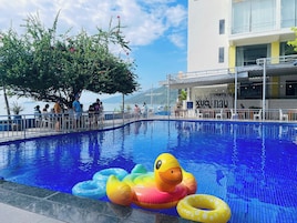 A pool area with inflatable toys, a dining area with tables and chairs, and a view of the sea.