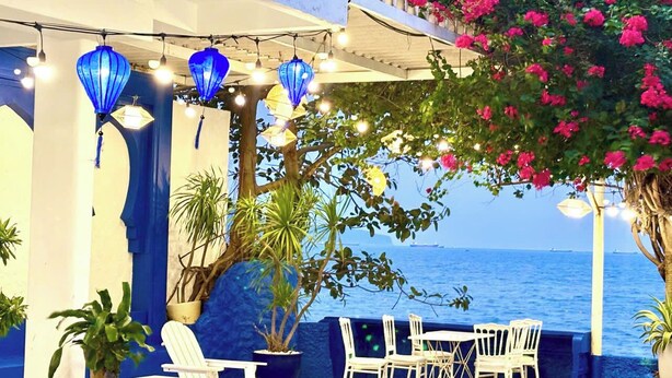 Un espace extérieur aménagé avec des chaises et des tables blanches, des lanternes bleues, des plantes en pot et une vue sur la mer.