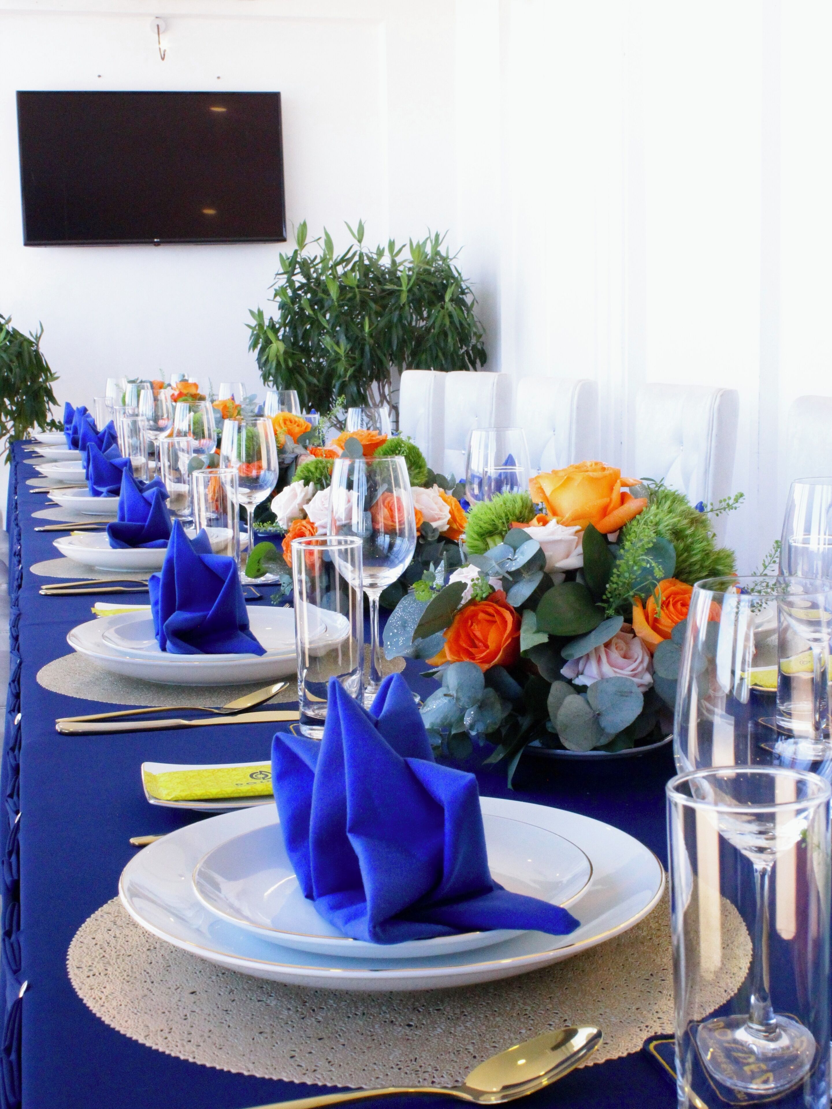 Una larga mesa de comedor con servilletas azules, vasos transparentes y centros de mesa con flores.