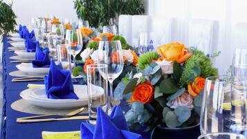 Una larga mesa de comedor con servilletas azules, vasos transparentes y centros de mesa con flores.