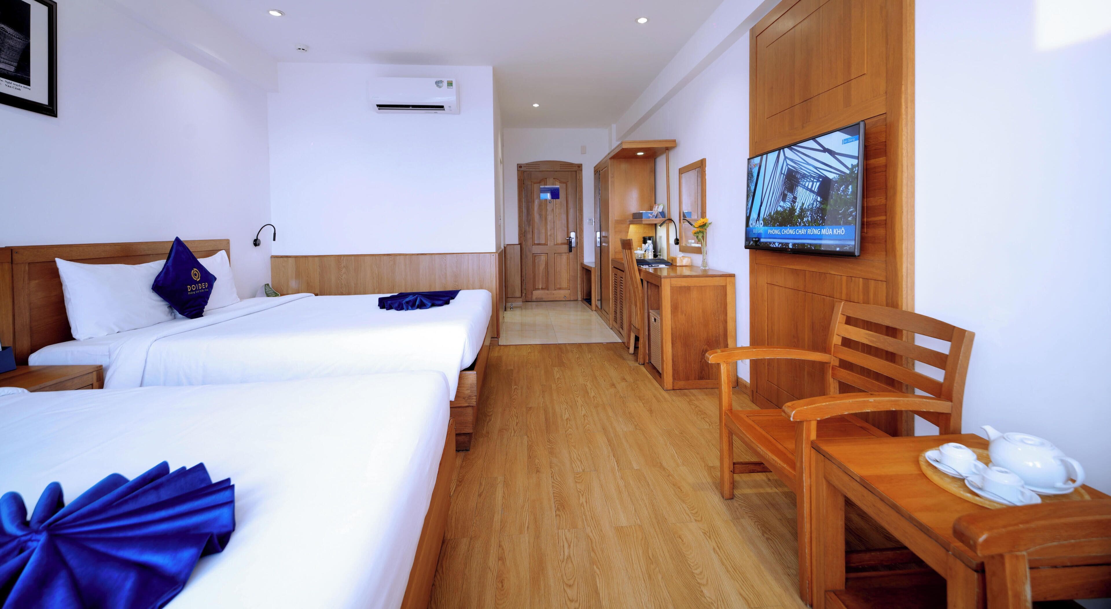 A hotel room with two beds, a wooden desk with a teapot, a television, and a small kitchenette.