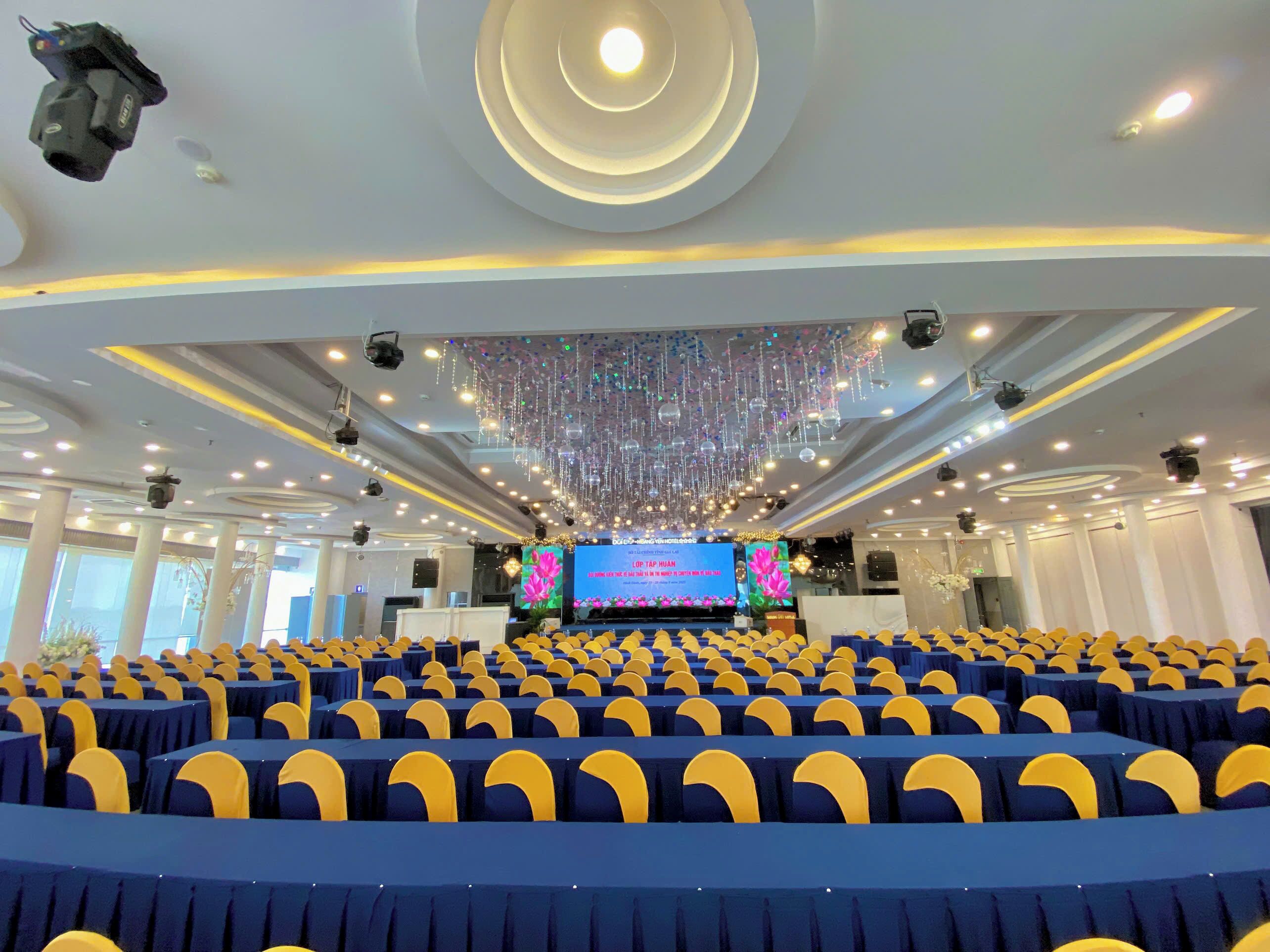 A conference room with rows of chairs, a stage with a large screen, and a ceiling with hanging lights and decorations.