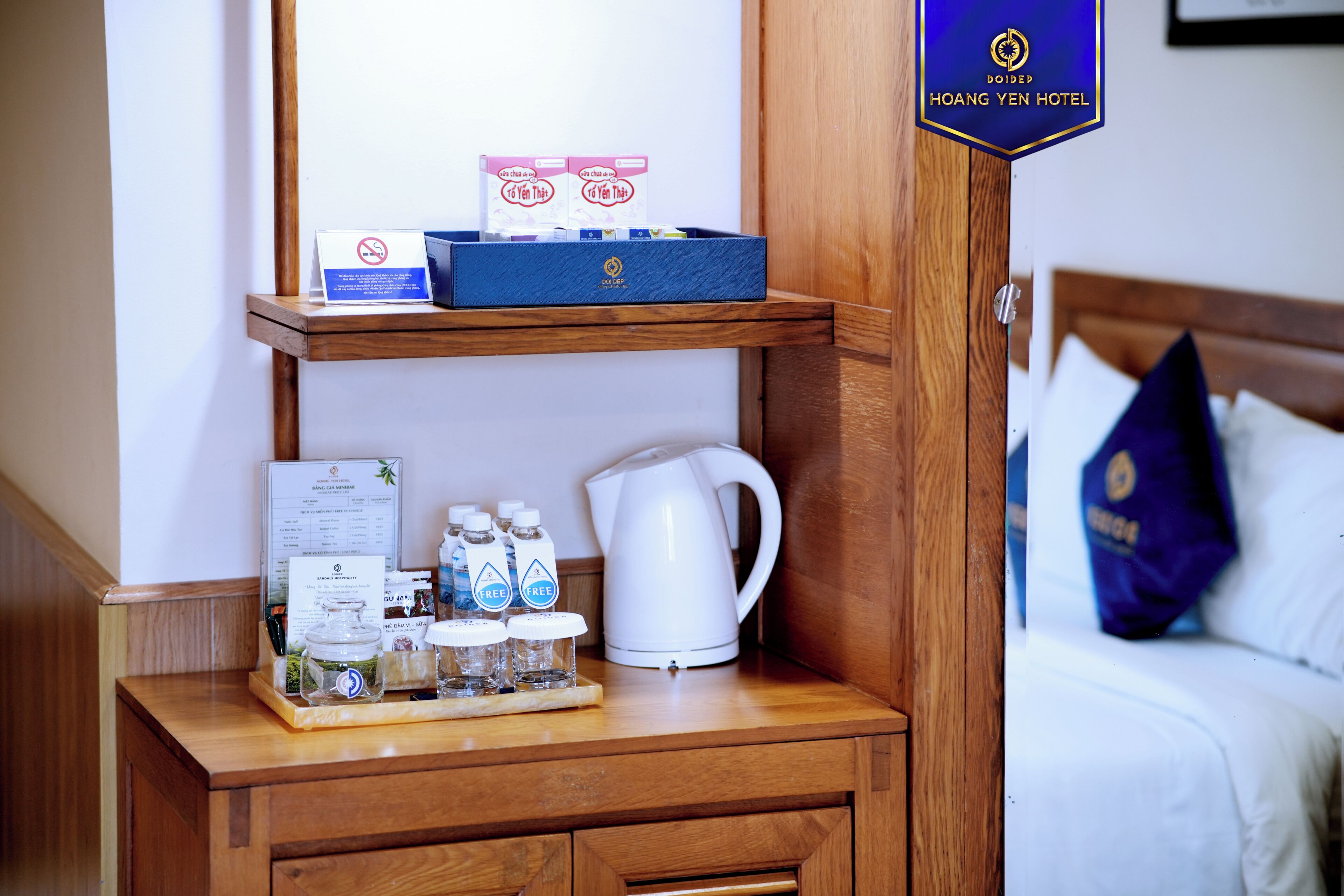 A hotel room with a wooden nightstand, a tray with bottled water, a white kettle, and a blue box with a sign.