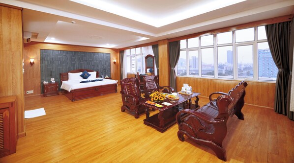 Une chambre d’hôtel avec un plancher en bois, un lit recouvert d’une courtepointe blanche, un bureau en bois orné d’un vase de fleurs et une vue sur la ville, visible à travers de grandes fenêtres.