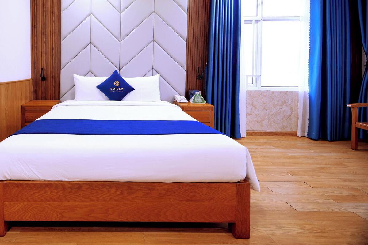 A hotel room with a wooden bed, a white headboard, a blue accent pillow, a wooden nightstand, and a window with blue curtains.