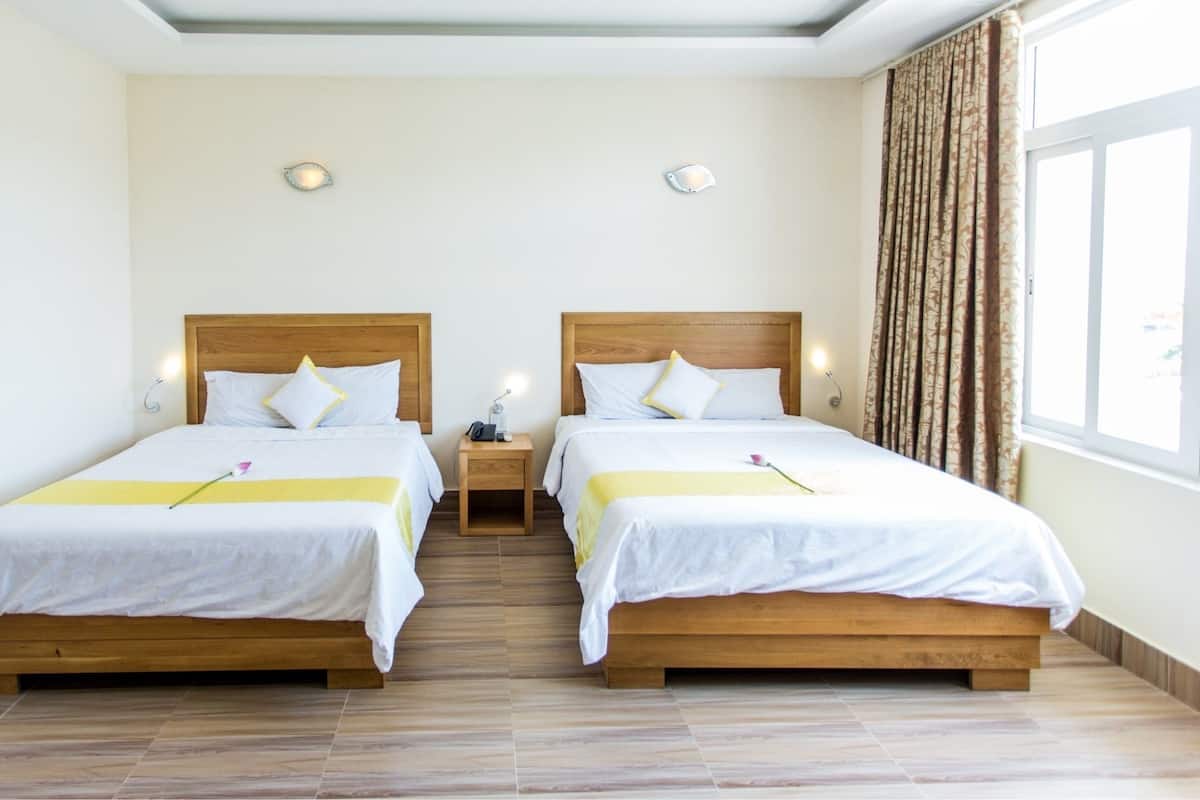 A hotel room with two beds, wooden headboards, white bedding, a bedside table with a lamp, and a window with curtains.