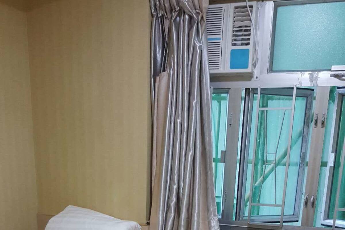 Quarto pequeno e limpo com uma cama de solteiro, uma janela com cortinas, uma pequena escrivaninha com um objeto rosa e um par de sapatos no chão.