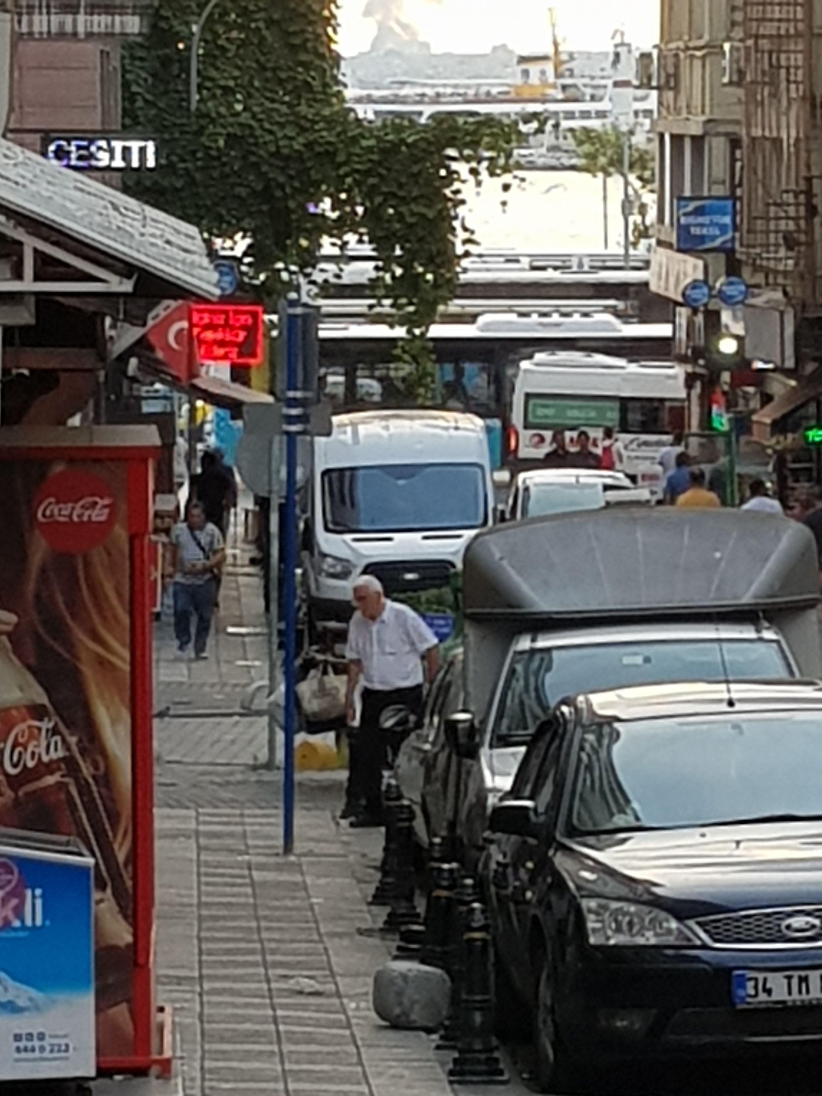 A busy street with parked cars, a Coca-Cola advertisement, and a sign for a Turkish restaurant.