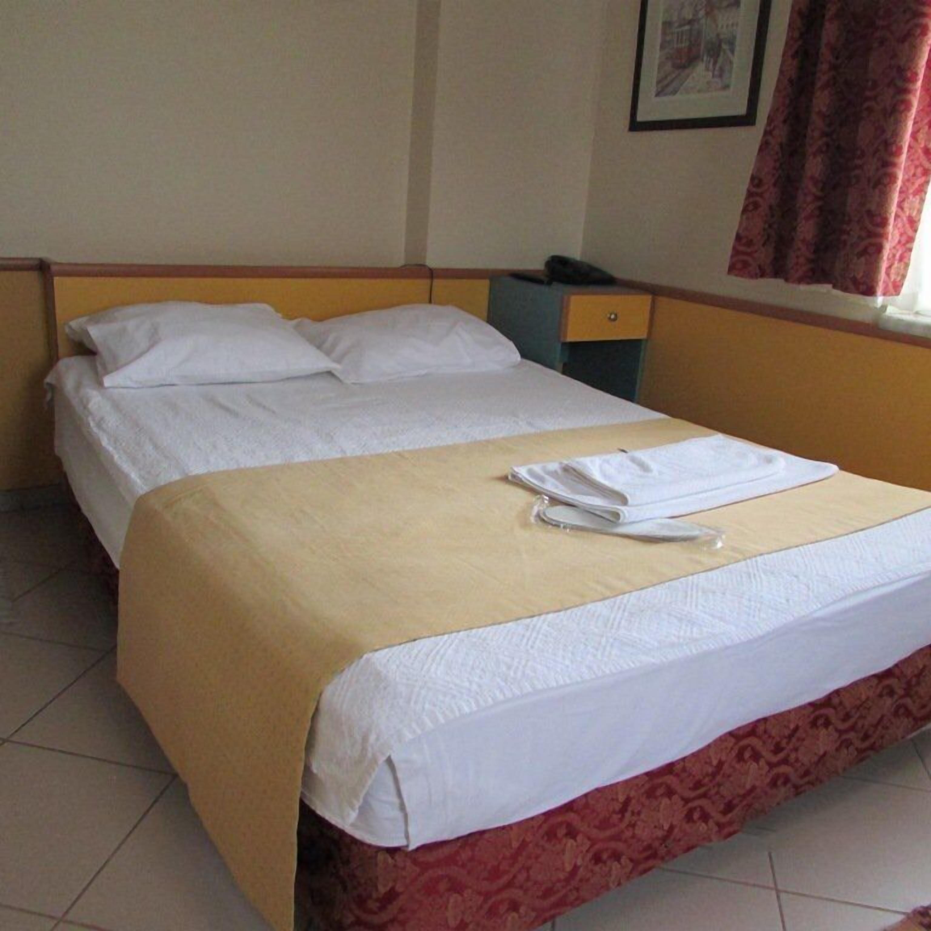A single bed with a beige bedspread and white linens, a small bedside table with a lamp, and a window with curtains.