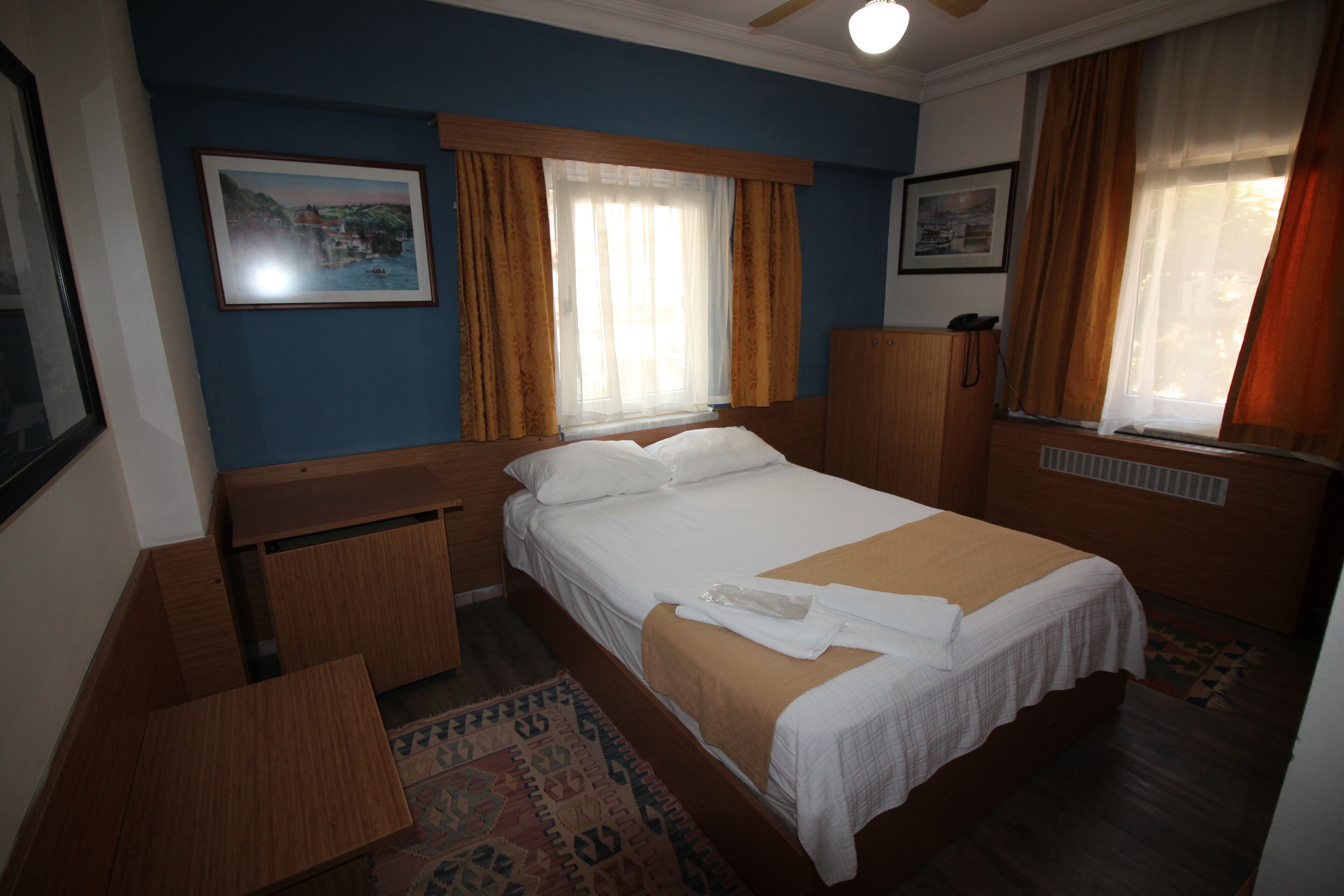 A hotel room with a bed, a wooden nightstand, a desk, a window with curtains, and a framed picture on the wall.