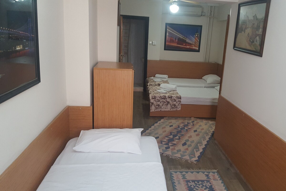 Habitación de hotel estrecha con una cama individual, un escritorio pequeño y un armario. Hay una alfombra estampada en el suelo y dos cuadros enmarcados en las paredes.