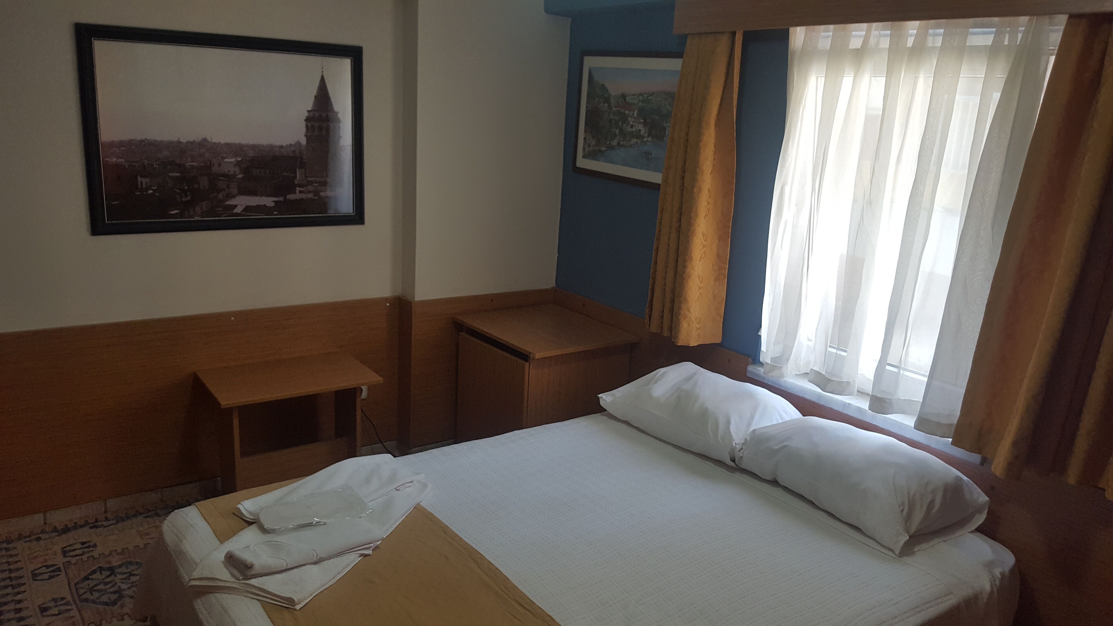 A hotel room with a bed, a wooden nightstand, a desk, and a window with curtains.