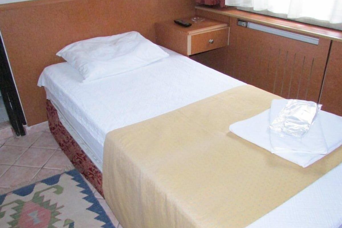 A hotel room with two single beds, a small bedside table with a remote control, a window with curtains, and a patterned rug.