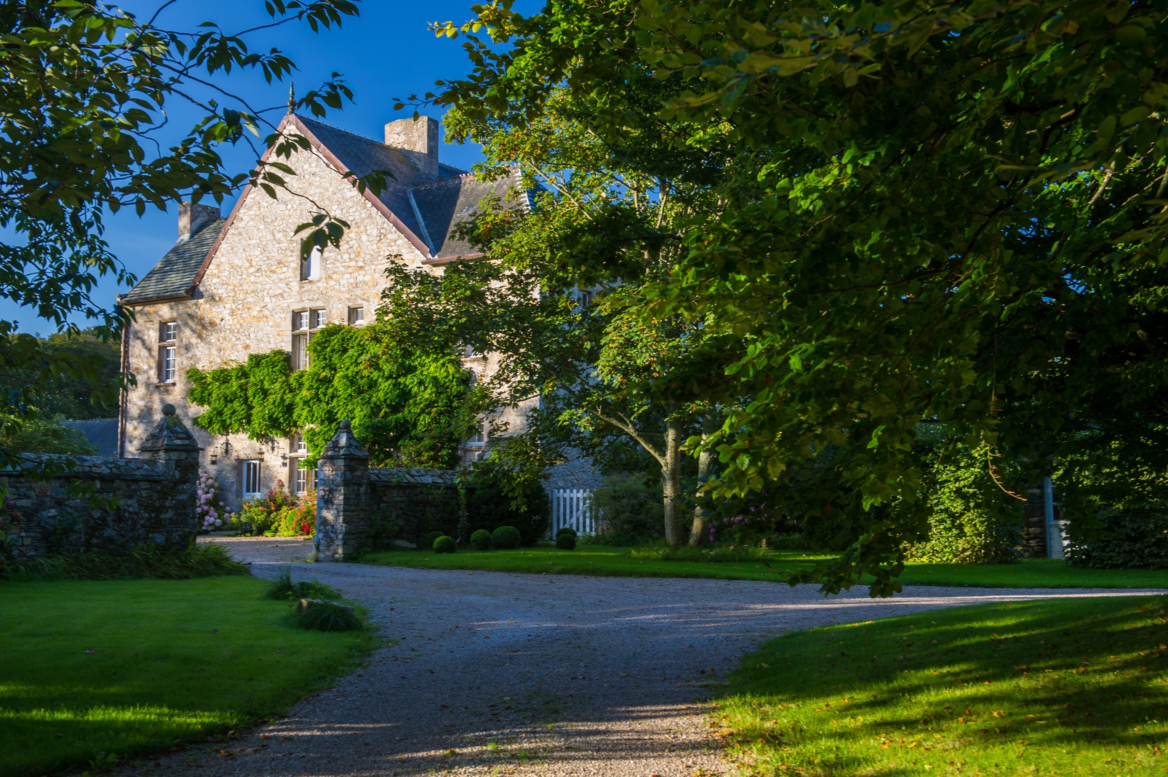 A stone building with a triangular roof, surrounded by lush greenery and a gravel driveway.