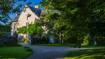 A stone building with a triangular roof, surrounded by lush greenery and a gravel driveway.