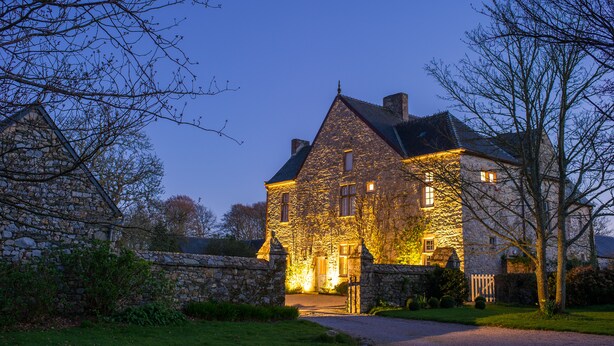 A stone house with a well-lit entrance, surrounded by a stone wall and a grassy area. - Le Manoir de la Fieffe (Cherbourg-en-Cotentin)