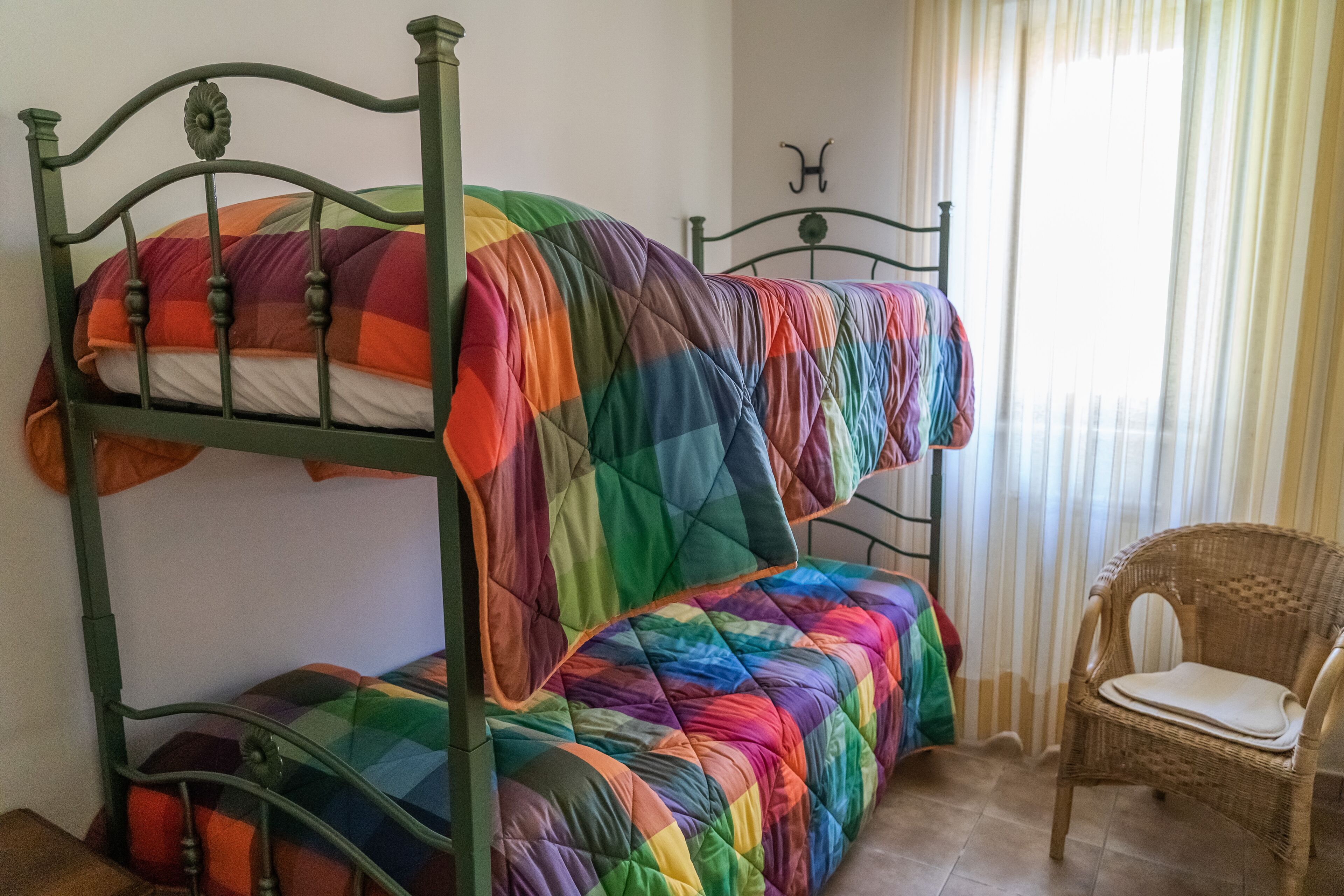 A bunk bed with a rainbow-colored quilt, a wicker chair, and a window with curtains.