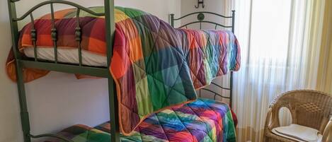 A bunk bed with a rainbow-colored quilt, a wicker chair, and a window with curtains.