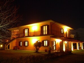 A two-story house with a balcony, multiple windows, and a well-lit exterior at night.