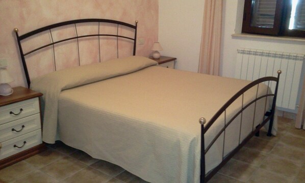 Una cama individual con estructura de madera y colchón color beige, una mesita de noche con lámpara y una ventana con persianas.